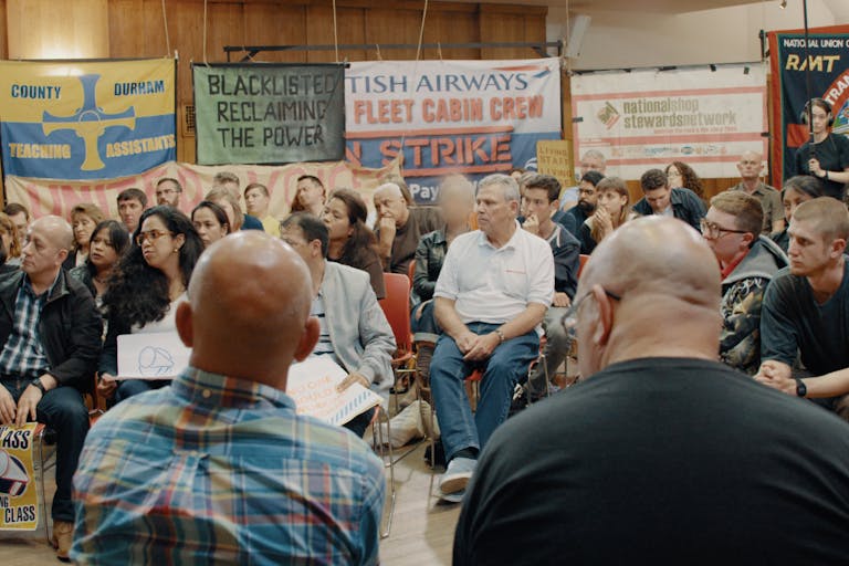 A room full of people having a community meeting with union and strike banners lining the room