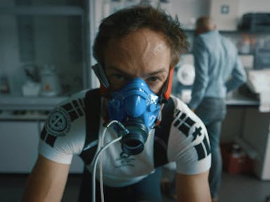 A white man in a blue fitness oxygen mask in cycling clothes riding a bike in a lab setting, looking strained