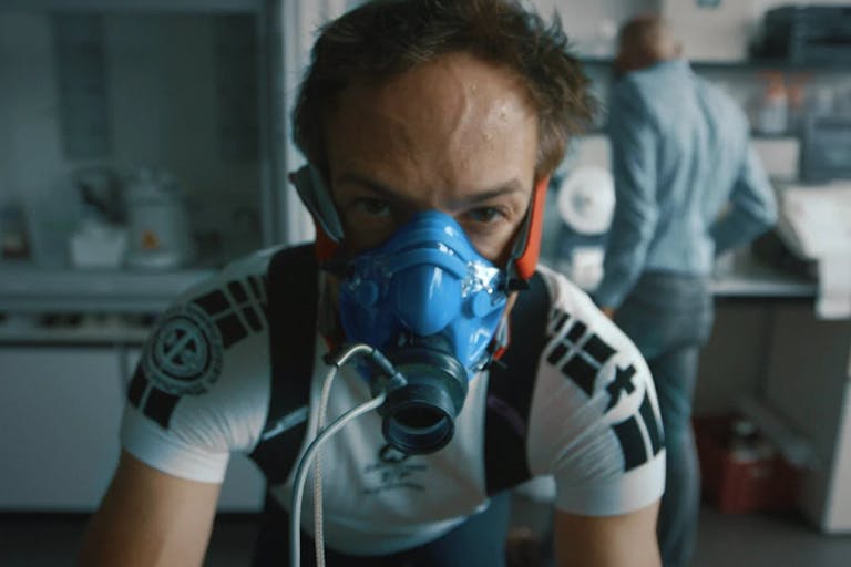 A white man in a blue fitness oxygen mask in cycling clothes riding a bike in a lab setting, looking strained