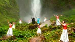 Two Indonesian men surrounded by Indonesian women in red tops and white skirts dancing in a lush green space in front of a waterfall