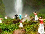 Two Indonesian men surrounded by Indonesian women in red tops and white skirts dancing in a lush green space in front of a waterfall