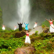 Two Indonesian men surrounded by Indonesian women in red tops and white skirts dancing in a lush green space in front of a waterfall