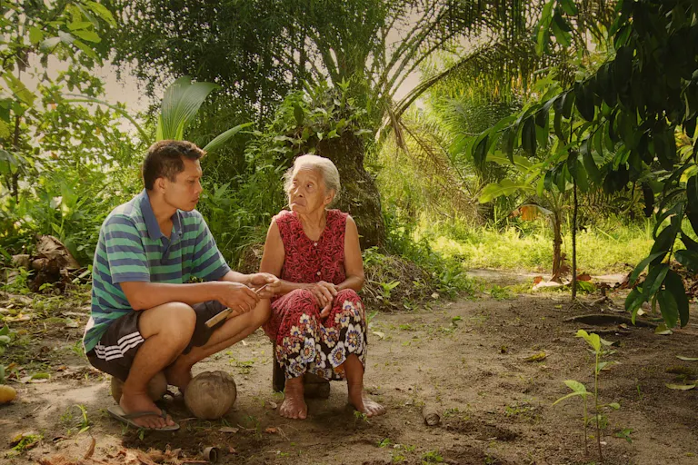 A young Indonesian man sits with an old Indonesian woman in a lush green forest