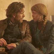 A white man with scruffy beard and a white woman with long ginger hair in a plait sitting on a sofa looking intently at each other