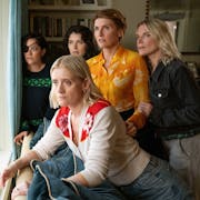 Five white women huddled in a living room looking out of a window suspiciously