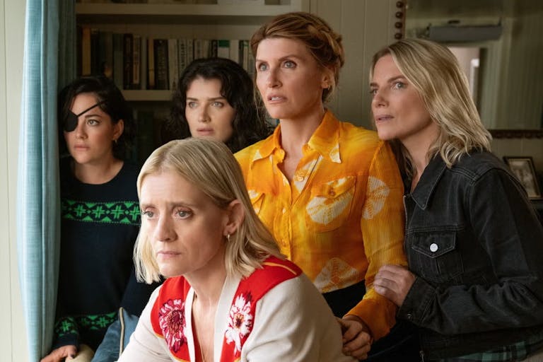 Five white women huddled in a living room looking out of a window suspiciously