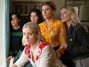 Five white women huddled in a living room looking out of a window suspiciously
