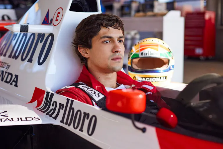 A young Brazilian man in a red jumpsuit sitting in a F1 car looking serious