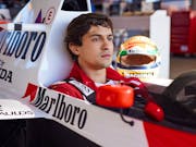 A young Brazilian man in a red jumpsuit sitting in a F1 car looking serious