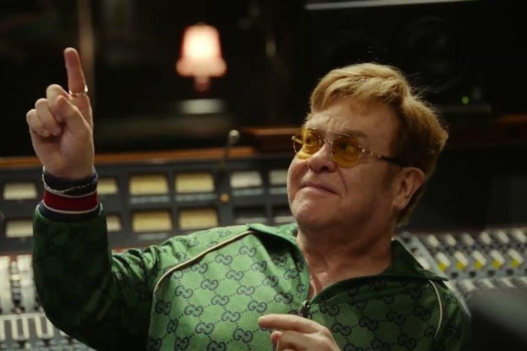 A middle aged white man in a green gucci tracksuit sitting in a recording studio smiling pointing upwards