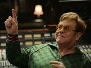 A middle aged white man in a green gucci tracksuit sitting in a recording studio smiling pointing upwards