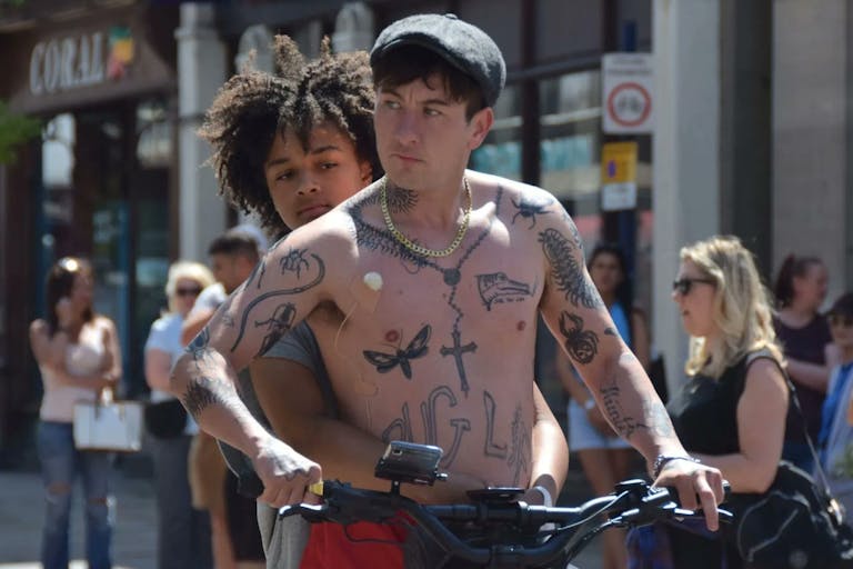 A young Black teenage girl holding onto a young white man with a chestful of tattoos as they ride an electric scooter