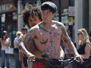 A young Black teenage girl holding onto a young white man with a chestful of tattoos as they ride an electric scooter