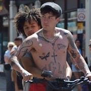 A young Black teenage girl holding onto a young white man with a chestful of tattoos as they ride an electric scooter