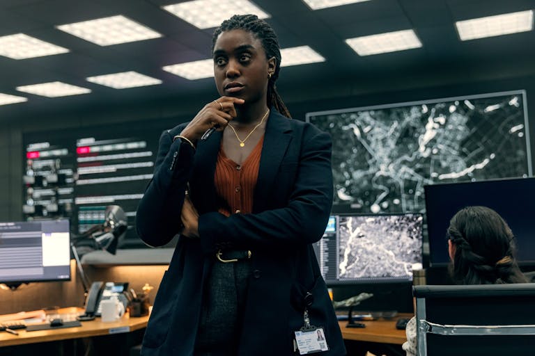 A Black woman in a suit leaning against a desk in an intelligence office with high tech computers and screens