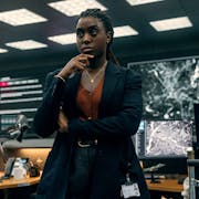A Black woman in a suit leaning against a desk in an intelligence office with high tech computers and screens