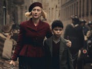 A young white woman walking down a war torn street in 1940s with her arm around a young mixed race Black boy