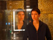 Young woman with short dark hair stands in front of a gold futuristic background, her image reflected in the glass beside her.