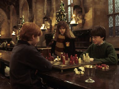 A young white girl and two young white boys sitting in a grand banquet hall with Christmas trees and flame lanterns playing table top chess