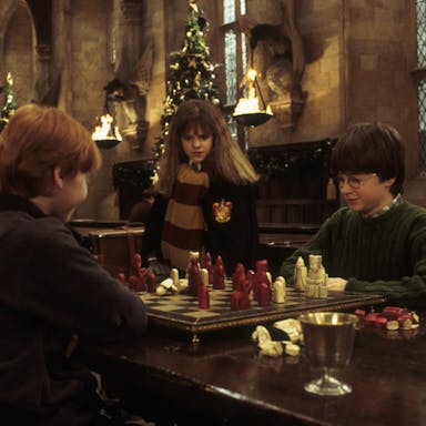 A young white girl and two young white boys sitting in a grand banquet hall with Christmas trees and flame lanterns playing table top chess
