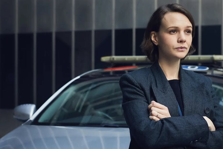 Young woman with short dark hair stands in front of a car with her arms folded, wearing a dark grey blazer jacket.