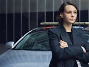 Young woman with short dark hair stands in front of a car with her arms folded, wearing a dark grey blazer jacket.
