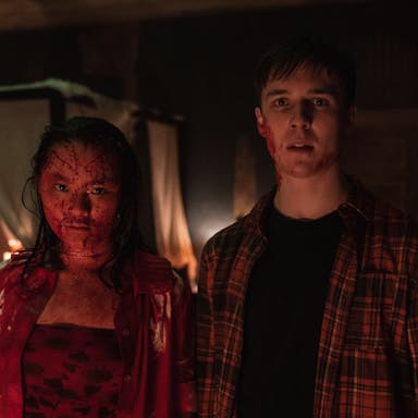 A young East Asian woman covered in blood standing next to a young white man looking shocked with a small amount of blood on his face