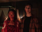 A young East Asian woman covered in blood standing next to a young white man looking shocked with a small amount of blood on his face