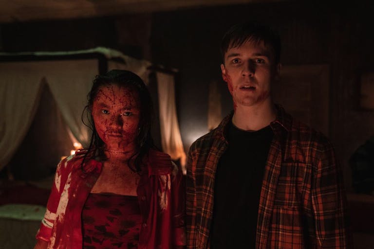 A young East Asian woman covered in blood standing next to a young white man looking shocked with a small amount of blood on his face