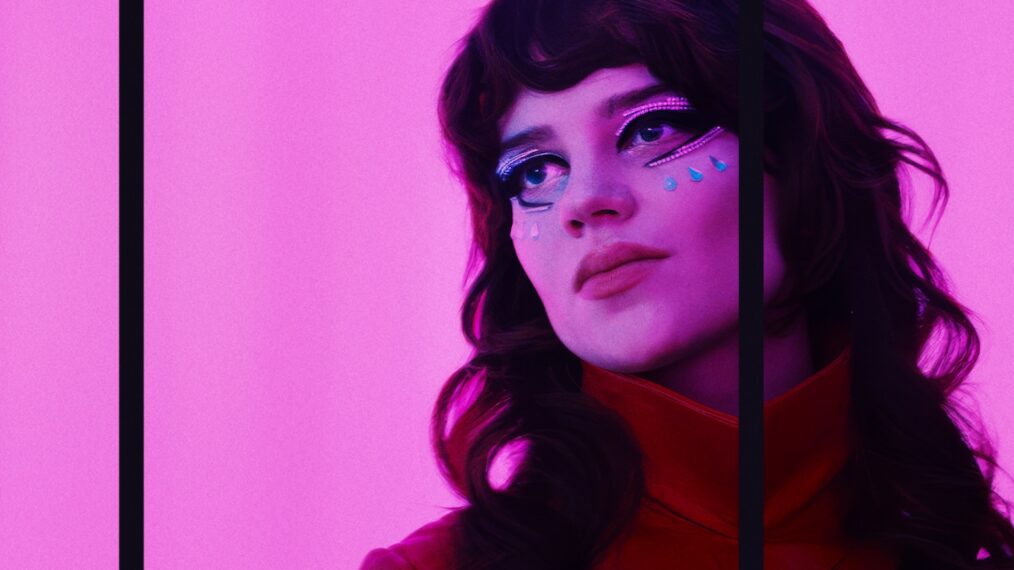 A close up of a dark-haired young white woman with face paint, bathed in a pink neon light