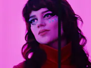 A close up of a dark-haired young white woman with face paint, bathed in a pink neon light