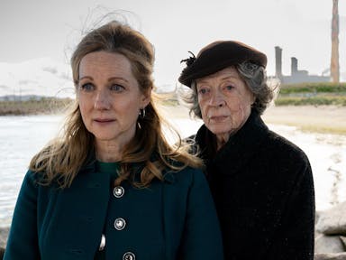 One middle aged woman and one old woman stand next to each other on a beach shore
