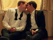 Two young white boys in dressy attire, sitting on a bedroom floor about to kiss