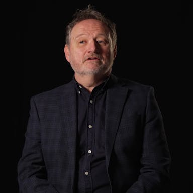 A middle aged white man in a dark suit and shirt, on screen name tag reads 'Neil Boyle, Director'