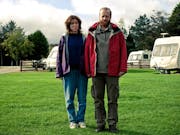 A white man and woman in outdoor clothing and waterproofs standing in a caravan park