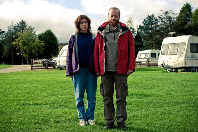 A white man and woman in outdoor clothing and waterproofs standing in a caravan park 