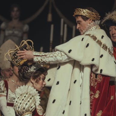 At a coronation, a white man in ostentatious royal regalia, a gold crown wreath on his head, places a large gold crown on a white woman's head also wearing royal clothing 