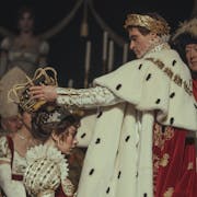 At a coronation, a white man in ostentatious royal regalia, a gold crown wreath on his head, places a large gold crown on a white woman's head also wearing royal clothing
