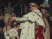 At a coronation, a white man in ostentatious royal regalia, a gold crown wreath on his head, places a large gold crown on a white woman's head also wearing royal clothing