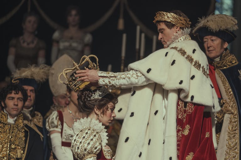 At a coronation, a white man in ostentatious royal regalia, a gold crown wreath on his head, places a large gold crown on a white woman's head also wearing royal clothing 