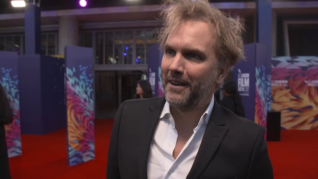 Man with grey hair talks on the red carpet of a film premiere.
