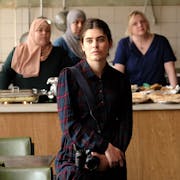 A young Syrian woman with a camera standing in a community kitchen space with other woman, some in head scarves.