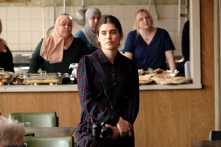 A young Syrian woman with a camera standing in a community kitchen space with other woman, some in head scarves. 