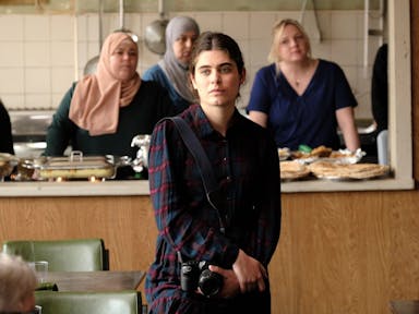 A young Syrian woman with a camera standing in a community kitchen space with other woman, some in head scarves. 