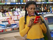 Young woman in a yellow sweater looks at her phone with a shocked expression, standing in a corner shop