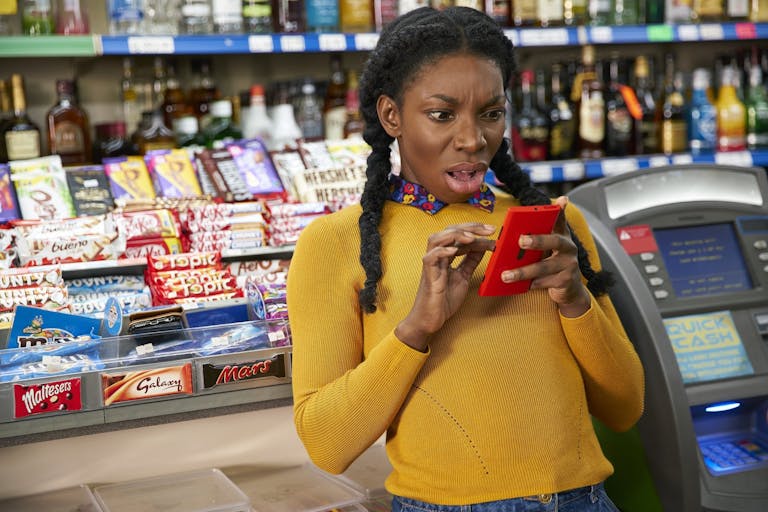 Young woman in a yellow sweater looks at her phone with a shocked expression, standing in a corner shop
