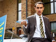 A young man in a suit wearing a diamond earring, sits on the bonnet of a car pouring out a bottle of water, with a yellow and blue 'To Let' sign beside him