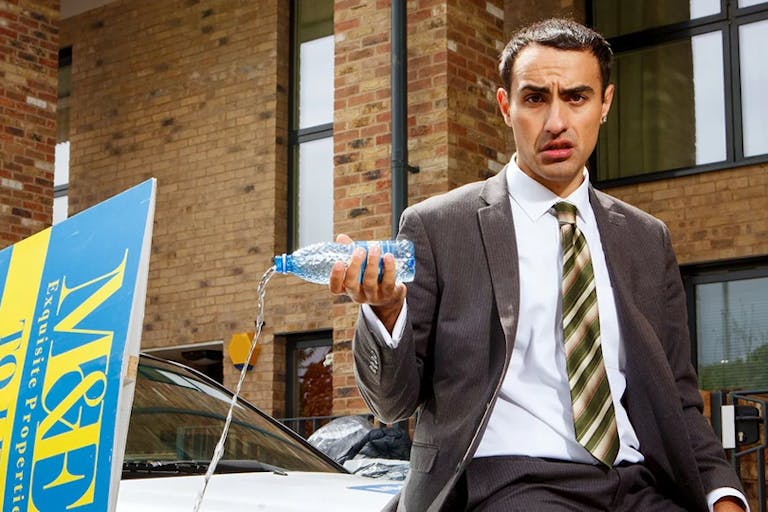 A young man in a suit wearing a diamond earring, sits on the bonnet of a car pouring out a bottle of water, with a yellow and blue 'To Let' sign beside him