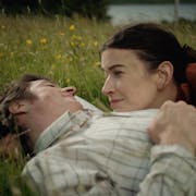 A white man and woman lay in a grassy meadow with buttercups surrounding them as they cuddle