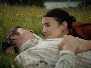 A white man and woman lay in a grassy meadow with buttercups surrounding them as they cuddle